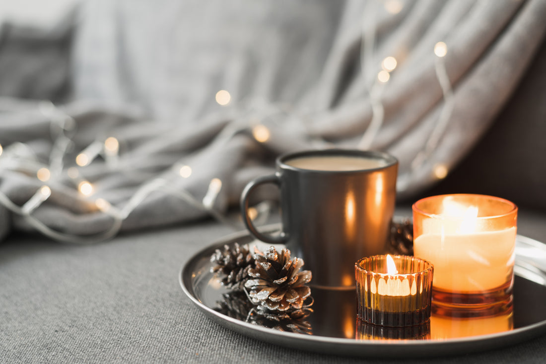 Two lit candles glowing on a round metal tray beside a black coffee mug and pine cones